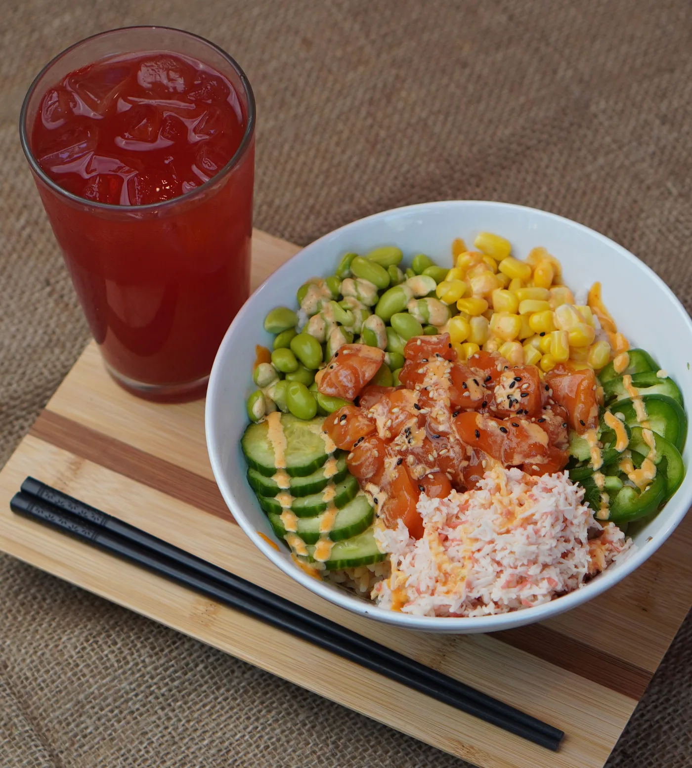A spicy salmon poke bowl with corn, edamame, cucumber and crab meat beside a red iced drink