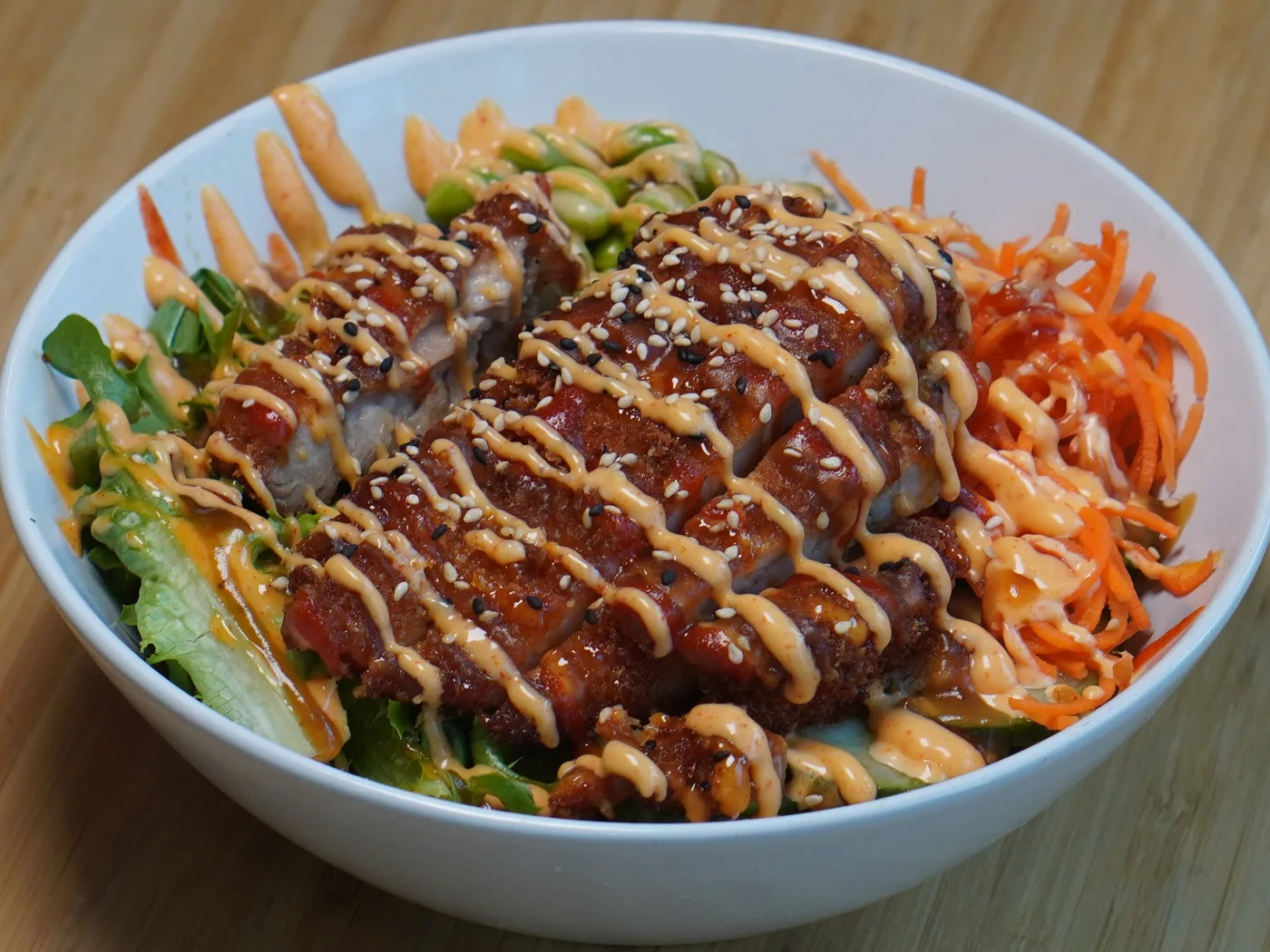 A crispy chicken bowl over mixed greens with carrots and edamame, drizzled with sauce