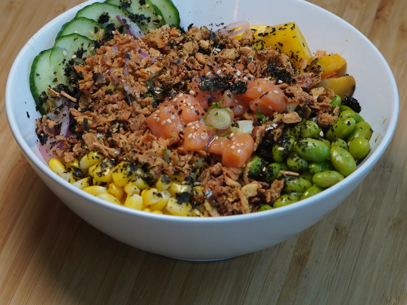 A poke bowl topped with salmon, crispy onions, corn, edamame, cucumber and mango